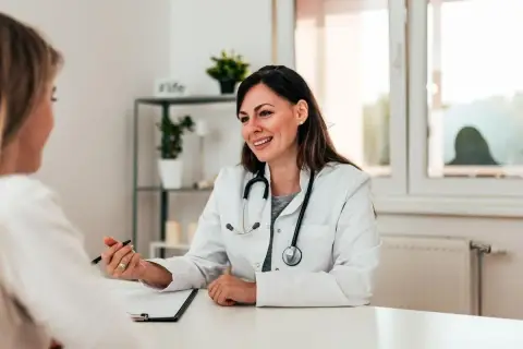 Medico sorridente parla con paziente, illustrando un punto con la penna. La scena evidenzia una buona comunicazione medico-paziente.