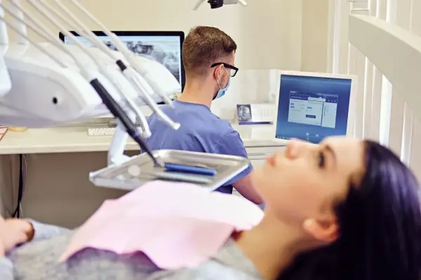 Un dentista con mascherina e occhiali lavora in uno studio dentistico moderno, esaminando radiografie su un monitor.
